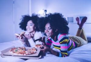 Women with afros wearing striped shirts eating pizza and watching TV