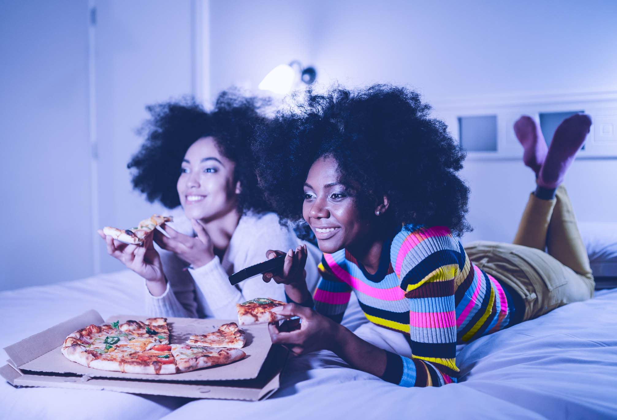 Women with afros wearing striped shirts eating pizza and watching TV
