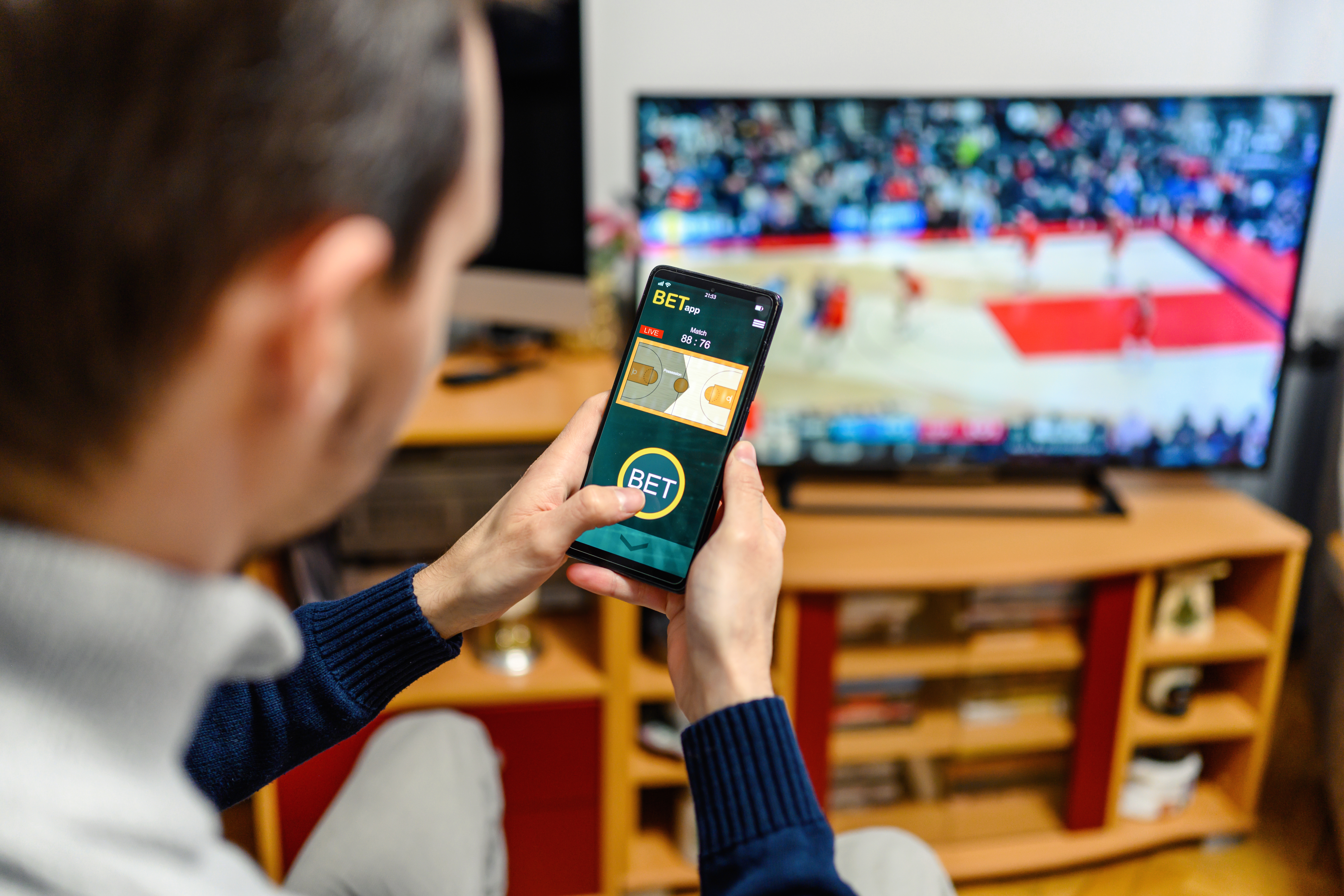 Young man sitting on the sofa at home and watching a basketball game and using a smartphone for sports betting.