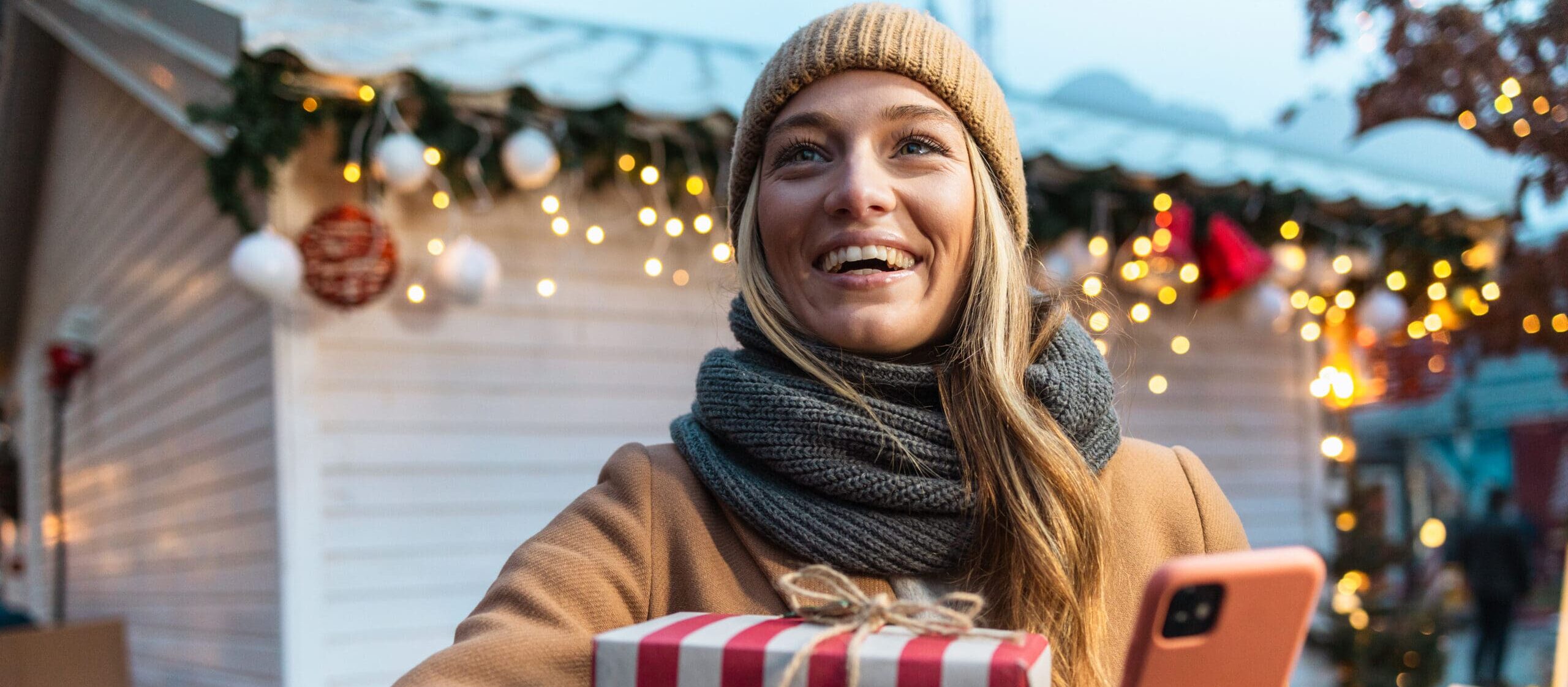 Excited young woman holding Christmas presents and using phone, on a Christmas market